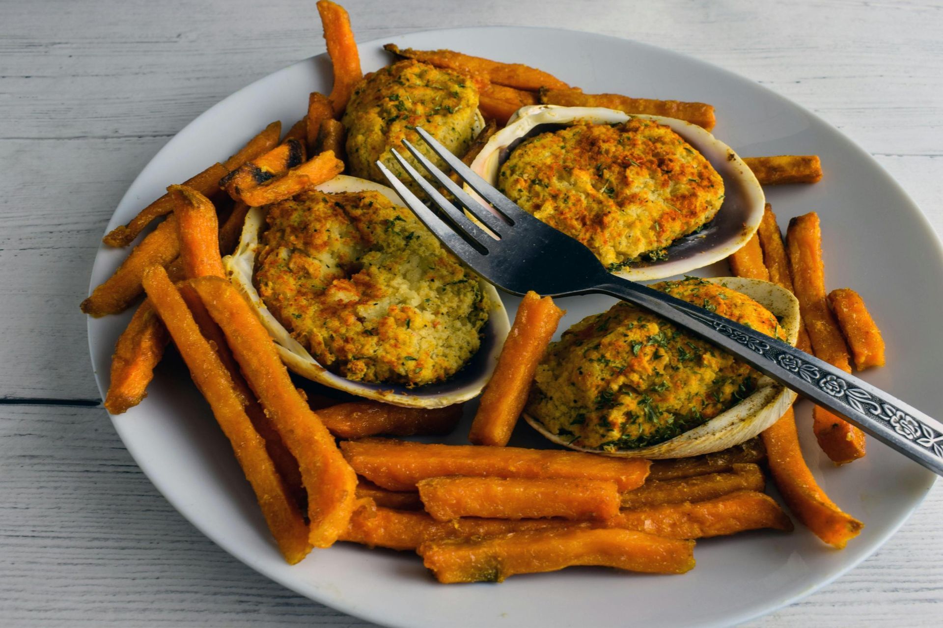 Three stuffed clam shells served alongside a pile of orange sweet potato fries on a white plate with a metal fork.