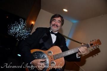 Man in a tuxedo smiling while playing an acoustic guitar at an indoor event
