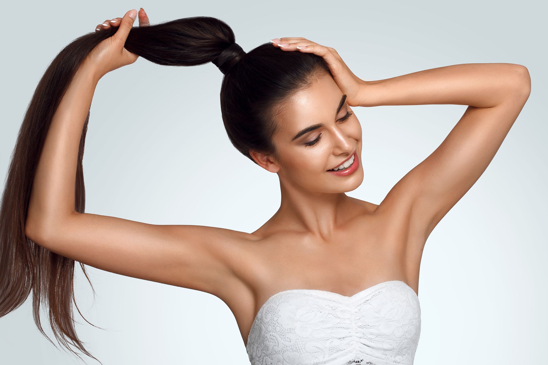 Woman with long brown hair, smiling, holding hair up in a high ponytail, wearing a white strapless top against a light blue background.