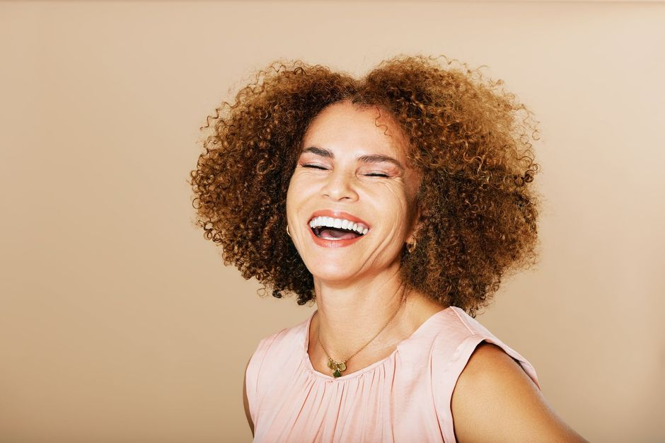 Woman with curly brown hair laughs, wearing a pink top, against a beige background.