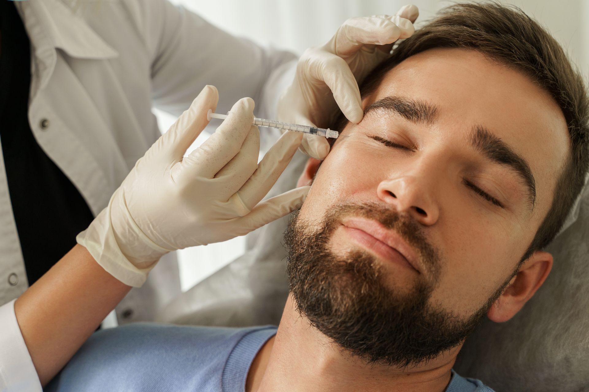A person receiving an injection near their eye. A gloved hand holds a syringe, and the person has their eyes closed.