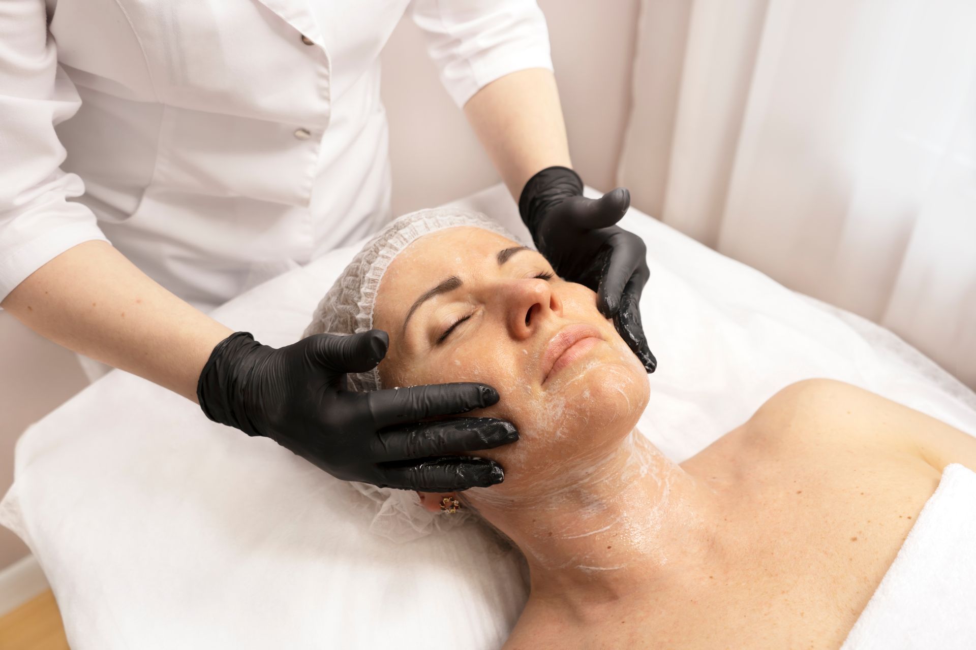 A person receiving a facial treatment. A gloved esthetician applies a scrub to the client's face and neck.
