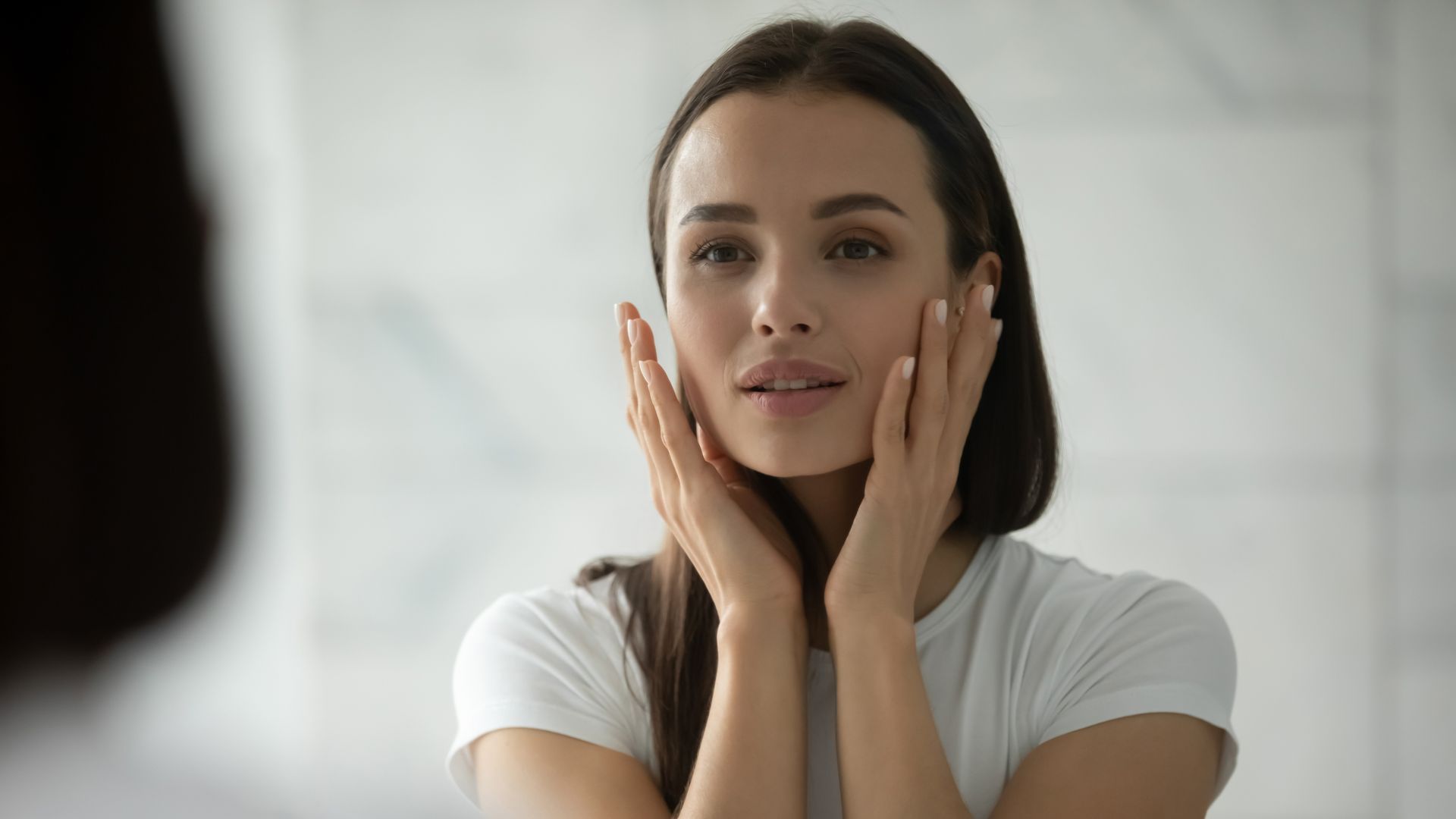 A woman with brown hair and a white tank top touches her forehead with a closed-eye expression, appearing thoughtful.