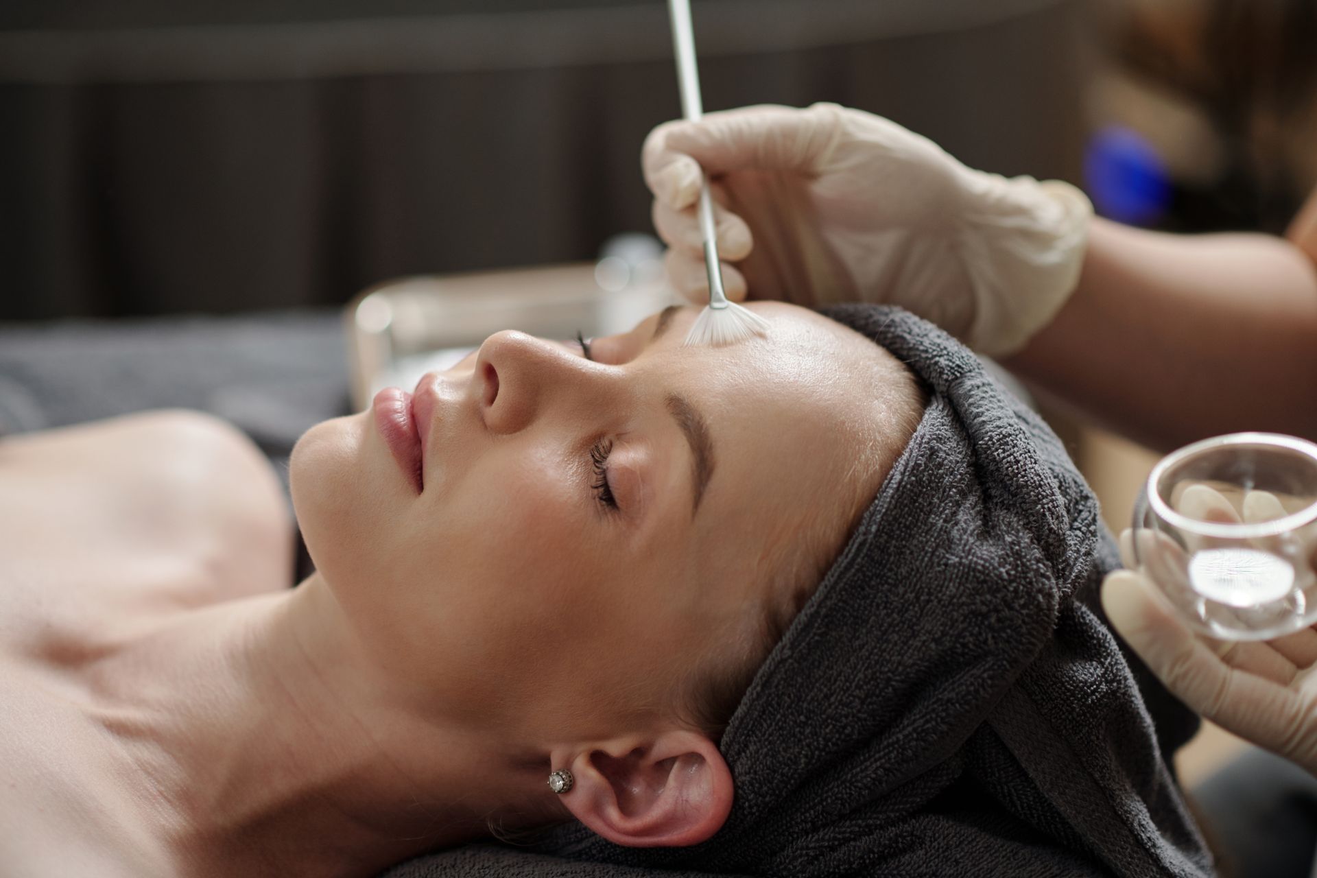 Woman receiving a facial treatment with a brush and glass bowl, lying with eyes closed under a towel wrap