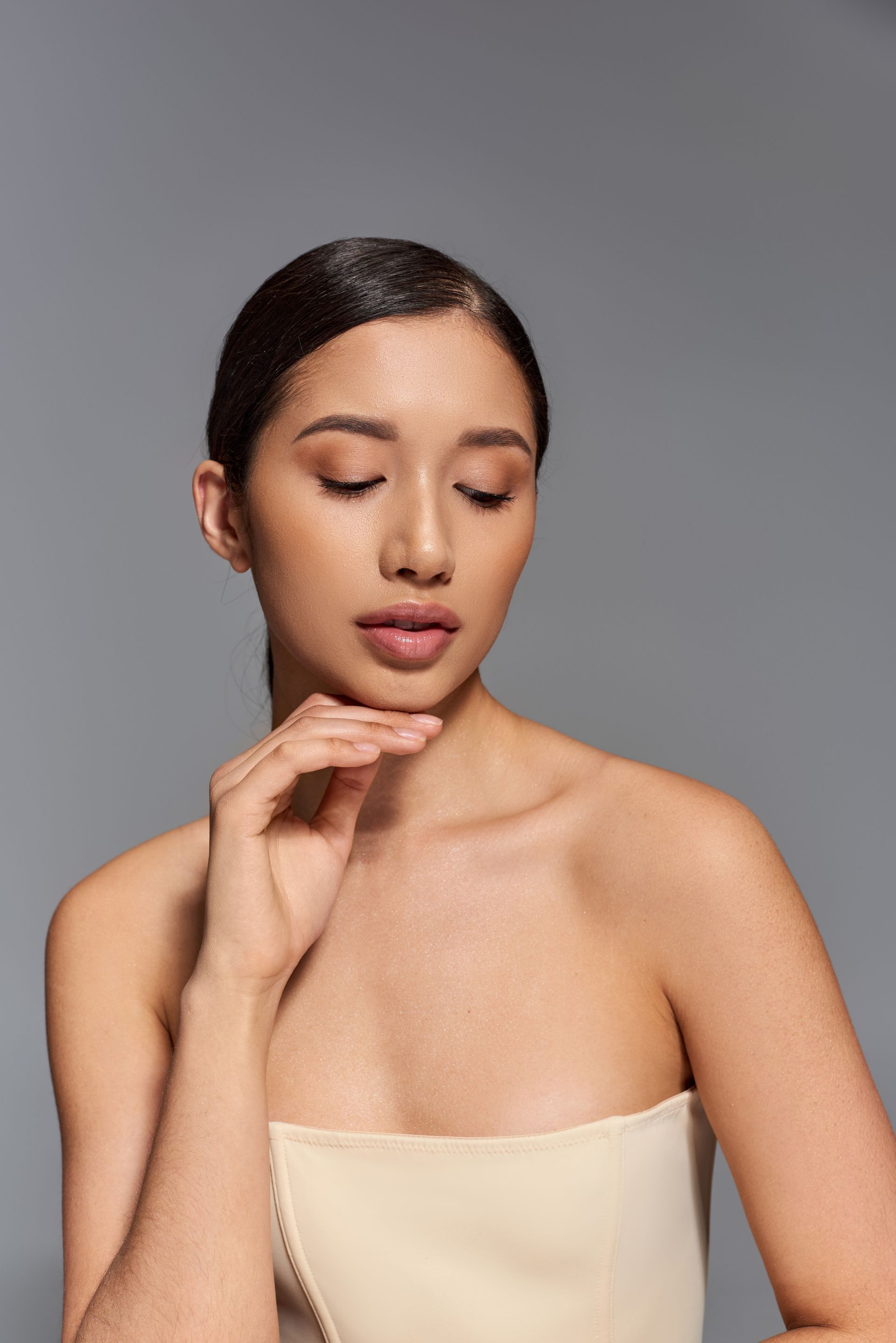 Woman with closed eyes and hand on chin, wearing a strapless beige top, against a gray background.