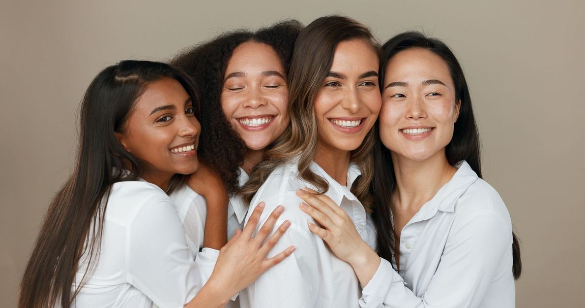 Four diverse women smiling and embracing, wearing white shirts, against a beige background.