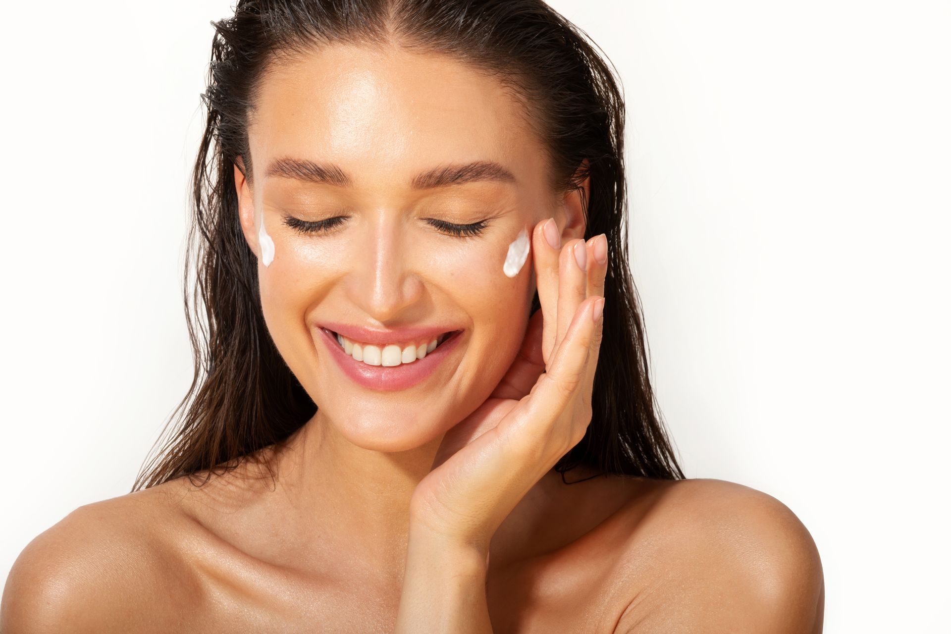 Smiling woman applying white cream to her cheek on a light background