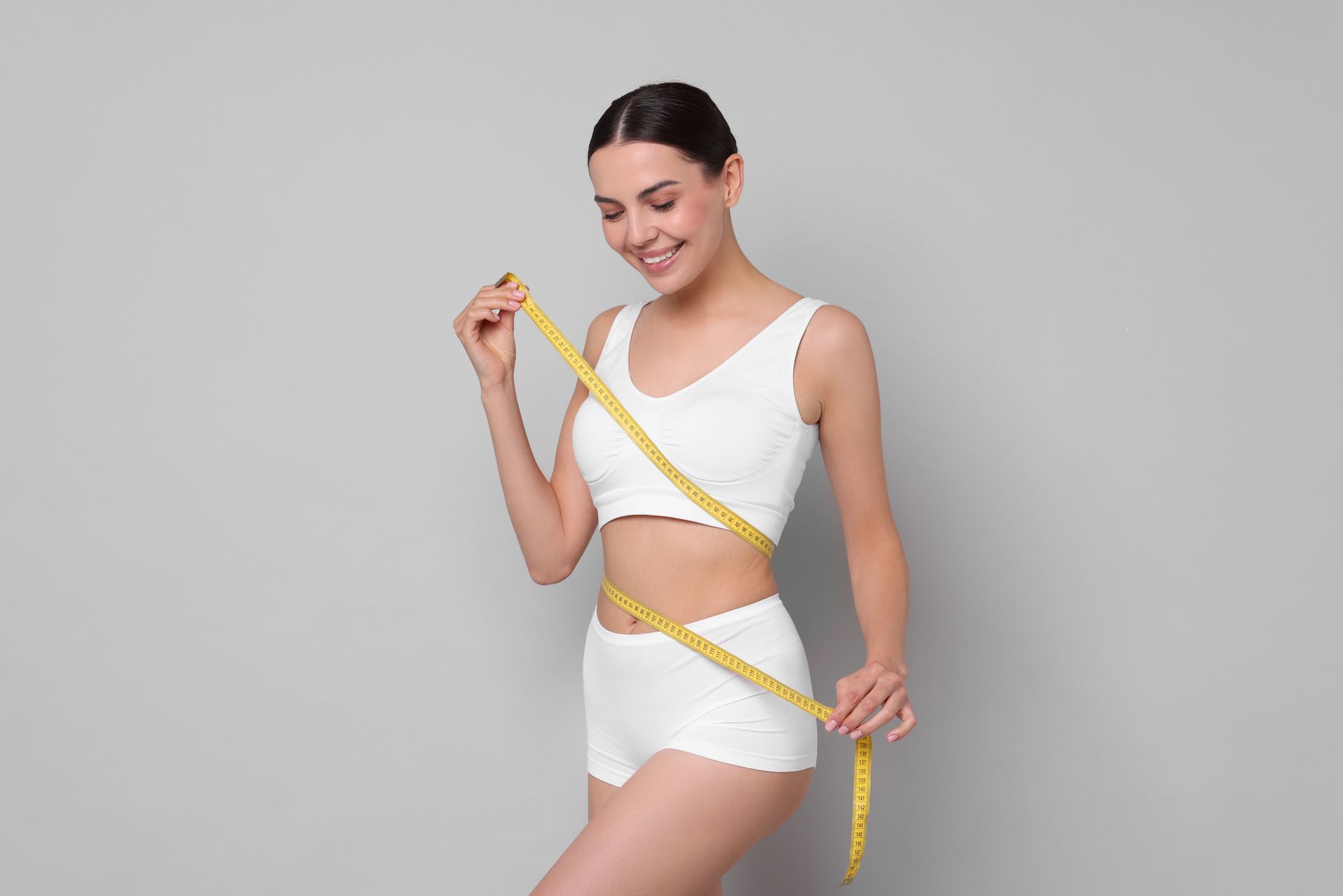 Woman measuring her waist with a yellow tape measure against a gray background