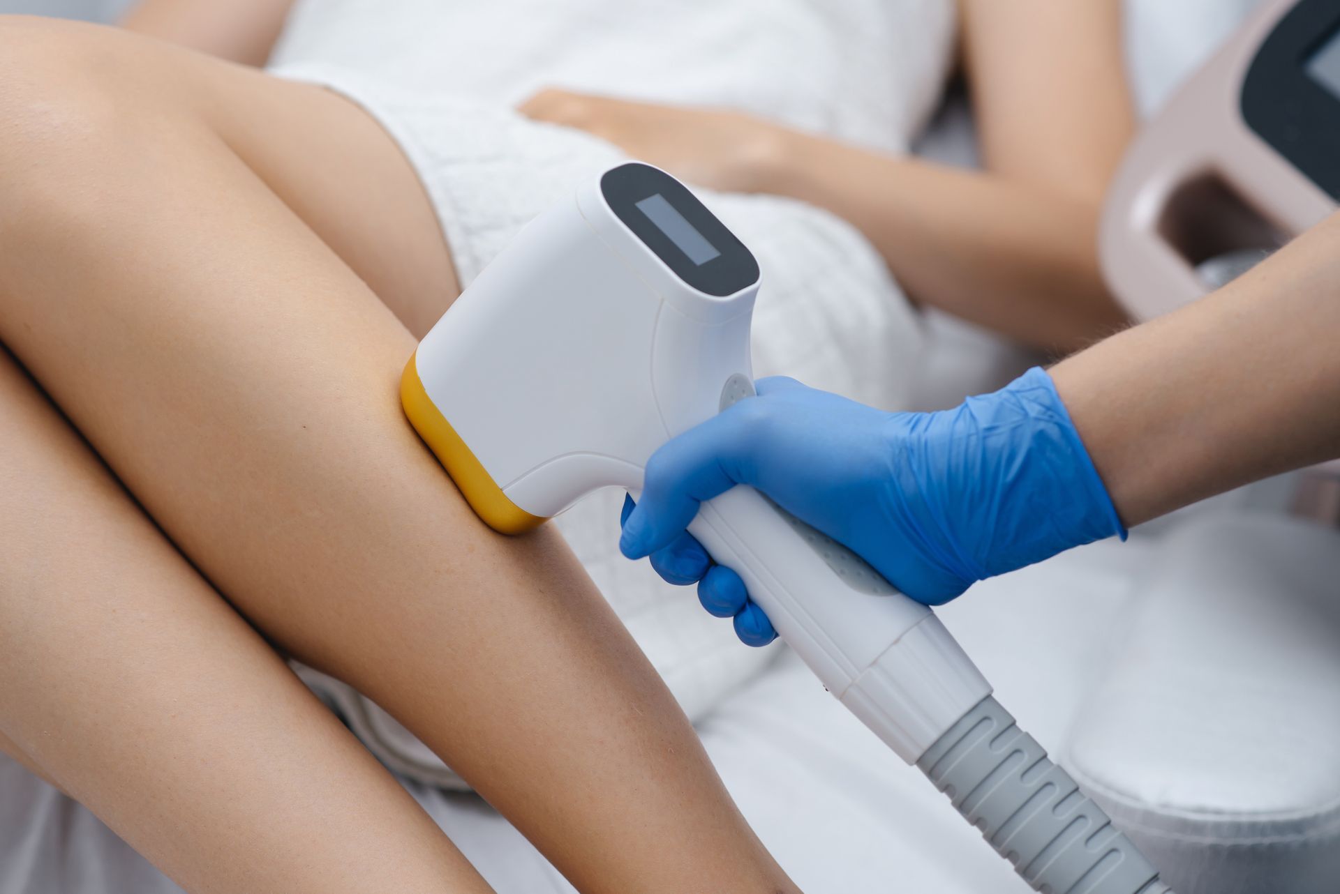 A person receiving laser hair removal on their leg. A technician in blue gloves holds the device.