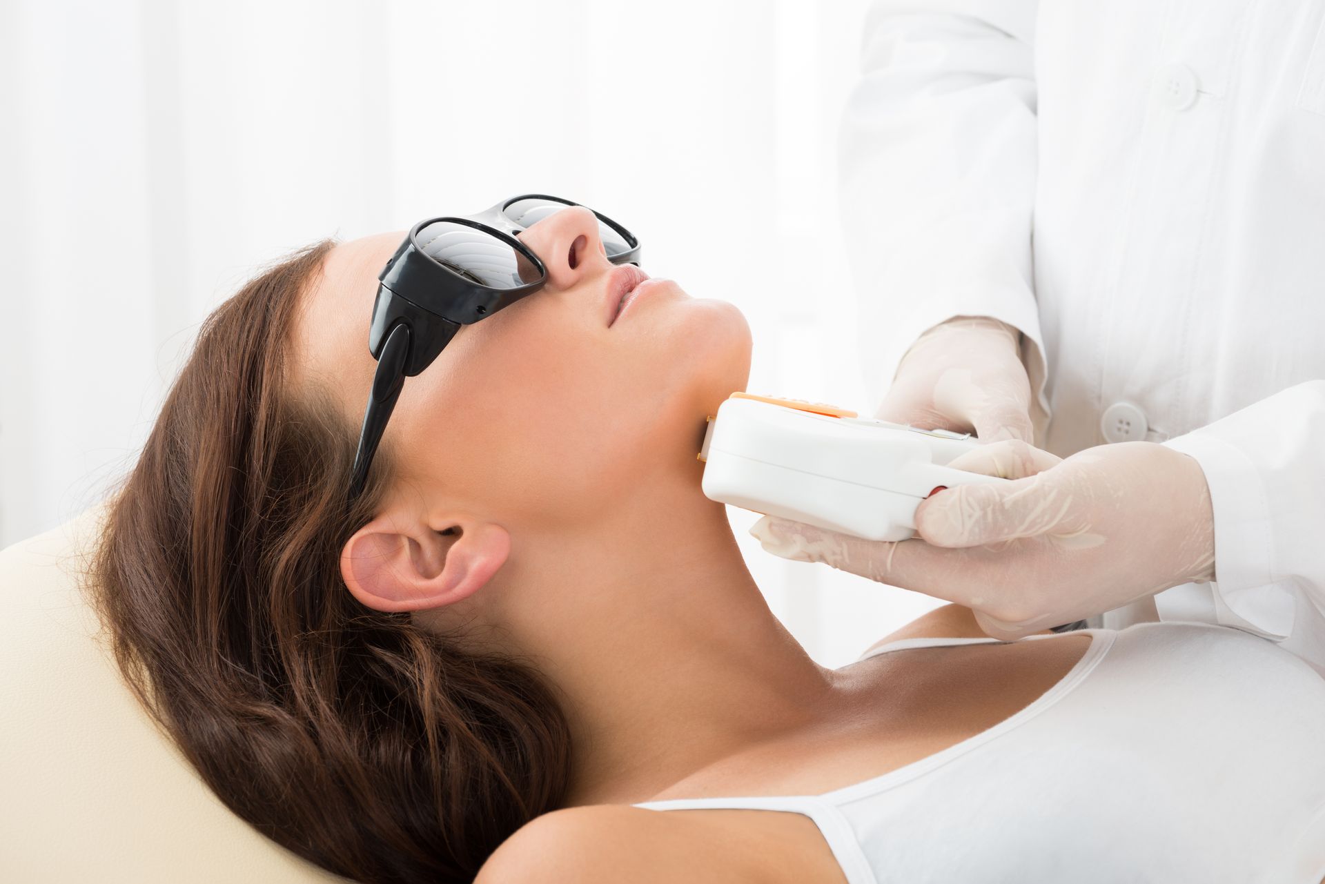 Woman receiving laser skin treatment on her neck from a gloved medical professional in a white setting. She wears protective eyewear.