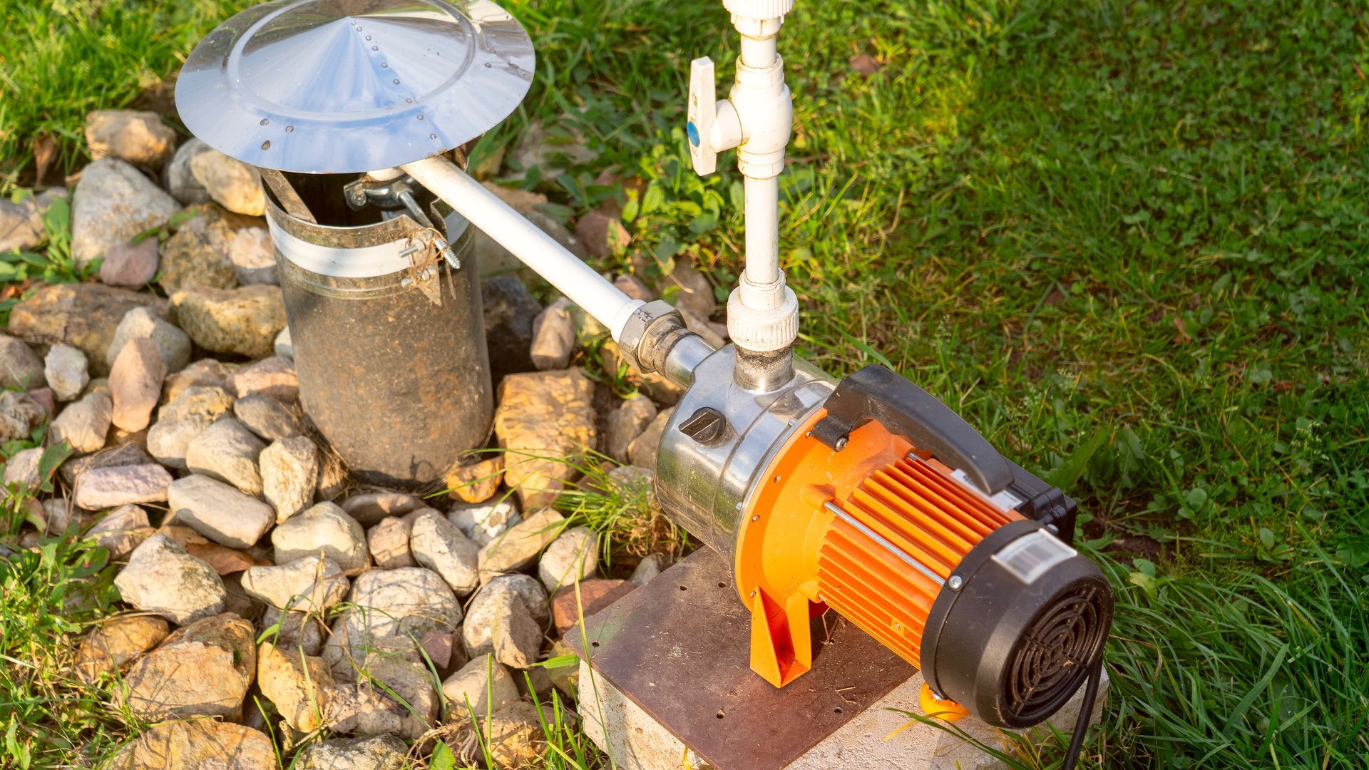 Small orange electric water pump connected to pipes beside rocks and grass outdoors