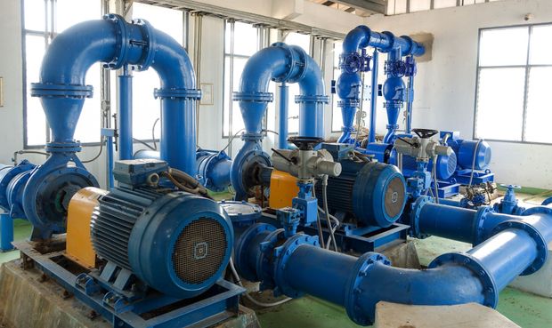 Blue industrial pumps and large pipes in a bright machinery room with windows.
