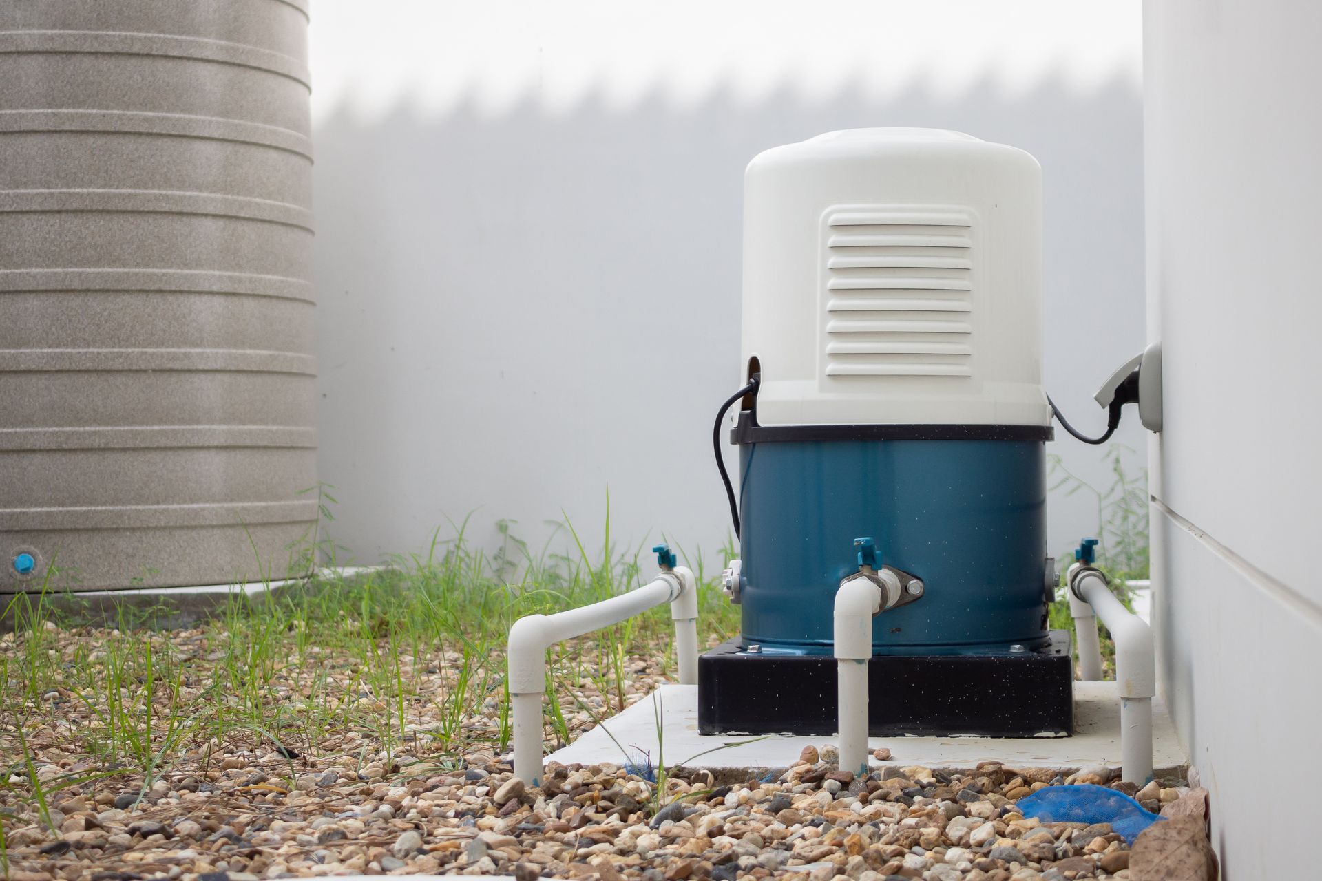 Outdoor water pump beside a corrugated tank, with white pipes on gravel and grass.
