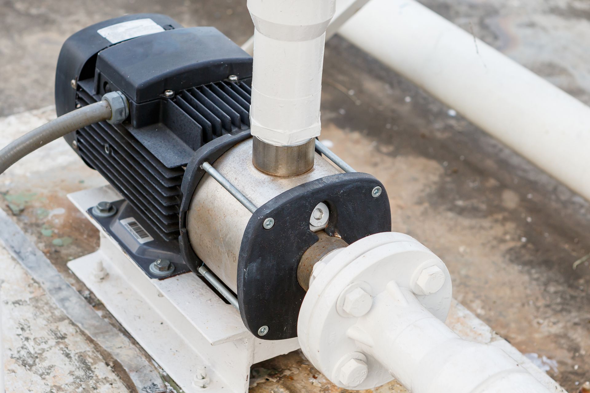 Close-up of a small electric water pump connected to white pipes on a concrete surface
