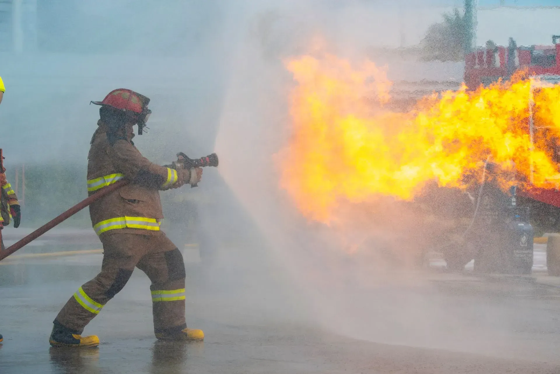 Firefighters spraying water at a large blaze beside a vehicle, with thick smoke and flames in the street.