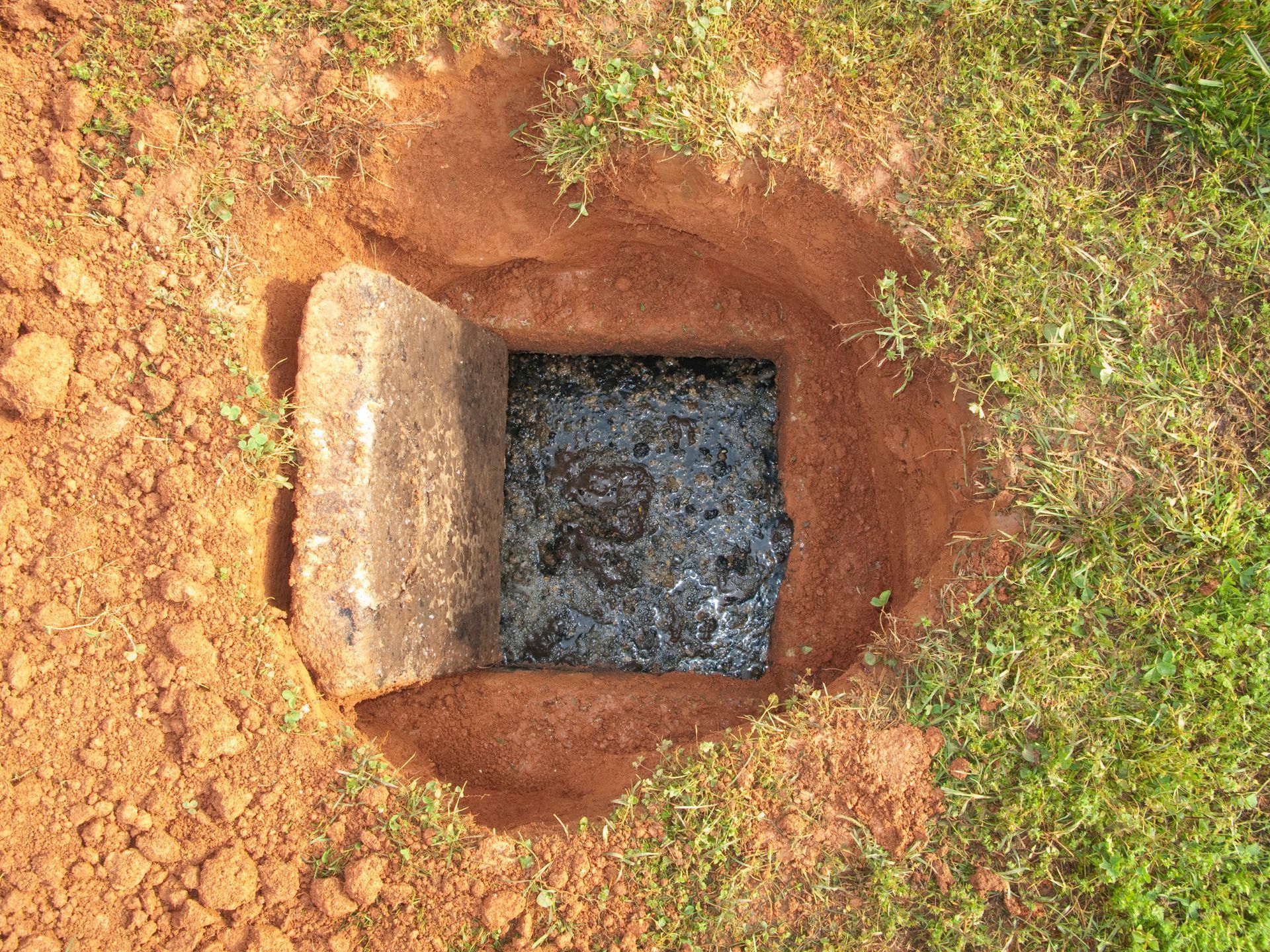 Open square pit with a concrete cover beside reddish soil and grass