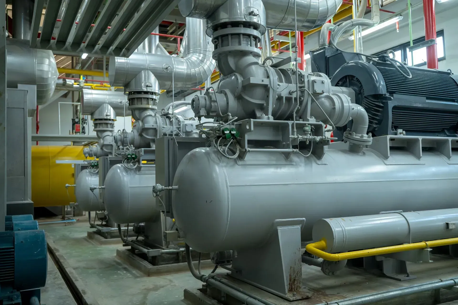 Industrial machinery and large gray tanks in a factory room with pipes and overhead ducts