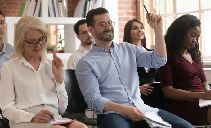 A group of people are sitting in a classroom and one of them is raising his hand to answer a question.