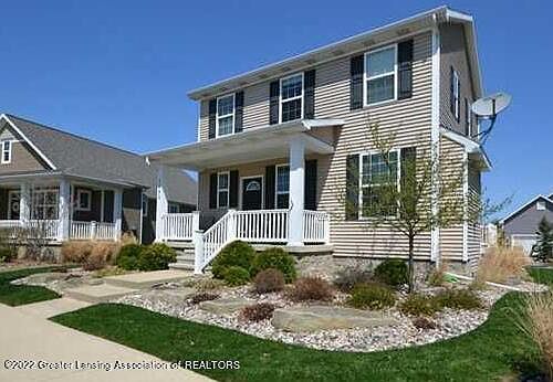 Two-story beige house with black shutters, porch, and landscaping under a blue sky.