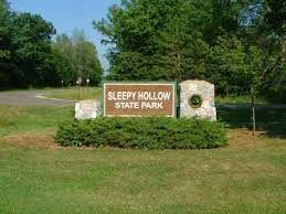 Sleepy Hollow State Park sign; brown sign, green bushes, stone pillars, grassy field, trees.