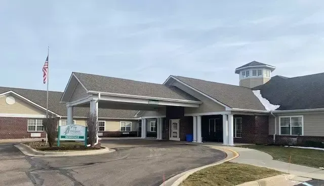 Exterior view of a beige and brick senior living facility with covered entrance, flagpole, and signage.