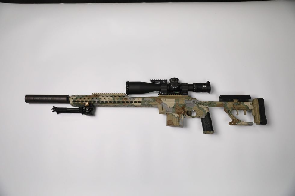 Camouflaged sniper rifle with scope, suppressor, and bipod against a white backdrop.