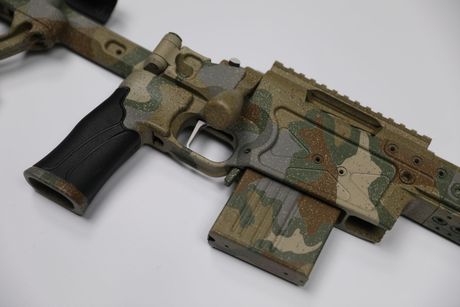 Camouflaged rifle with a black grip and magazine, on a white surface.