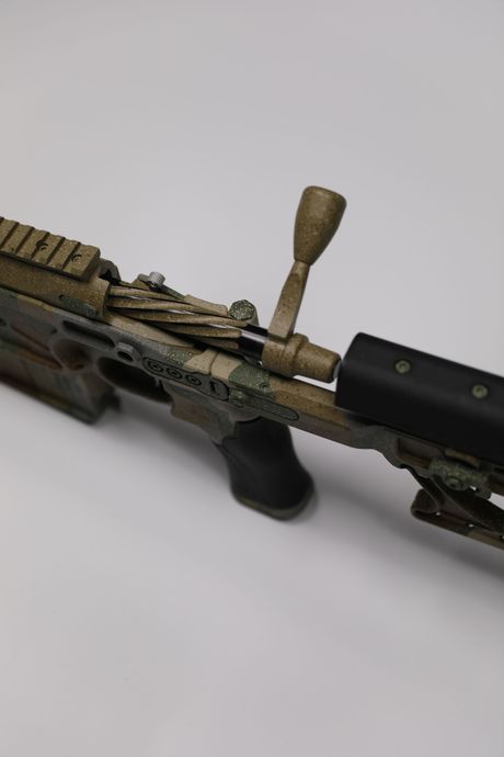Camouflaged bolt-action rifle, viewed from the side, resting on a white surface.