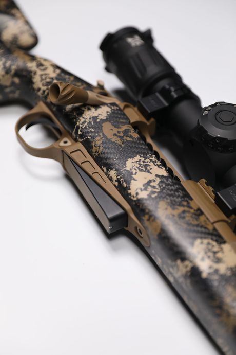 Close-up of a camouflage rifle with a scope, against a white backdrop.
