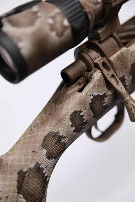 Bolt-action rifle with a camouflage pattern. Scope is mounted, and the rifle appears to be in a shooting position.
