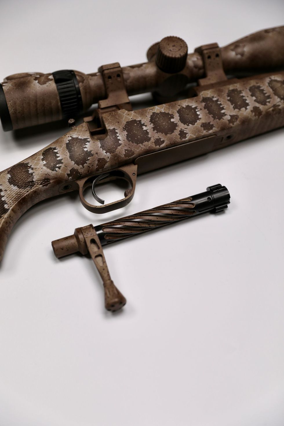 Brown and tan rifle with scope, bolt action disassembled, on white surface. Camouflage pattern.