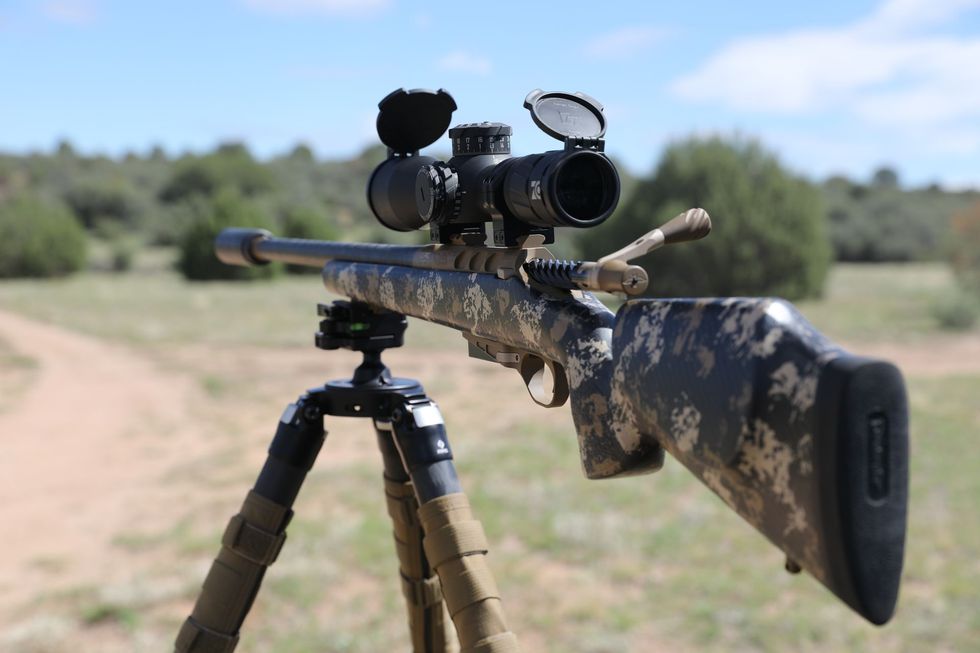 Rifle with camouflage pattern stock, mounted on a tripod, in a field, under a blue sky.