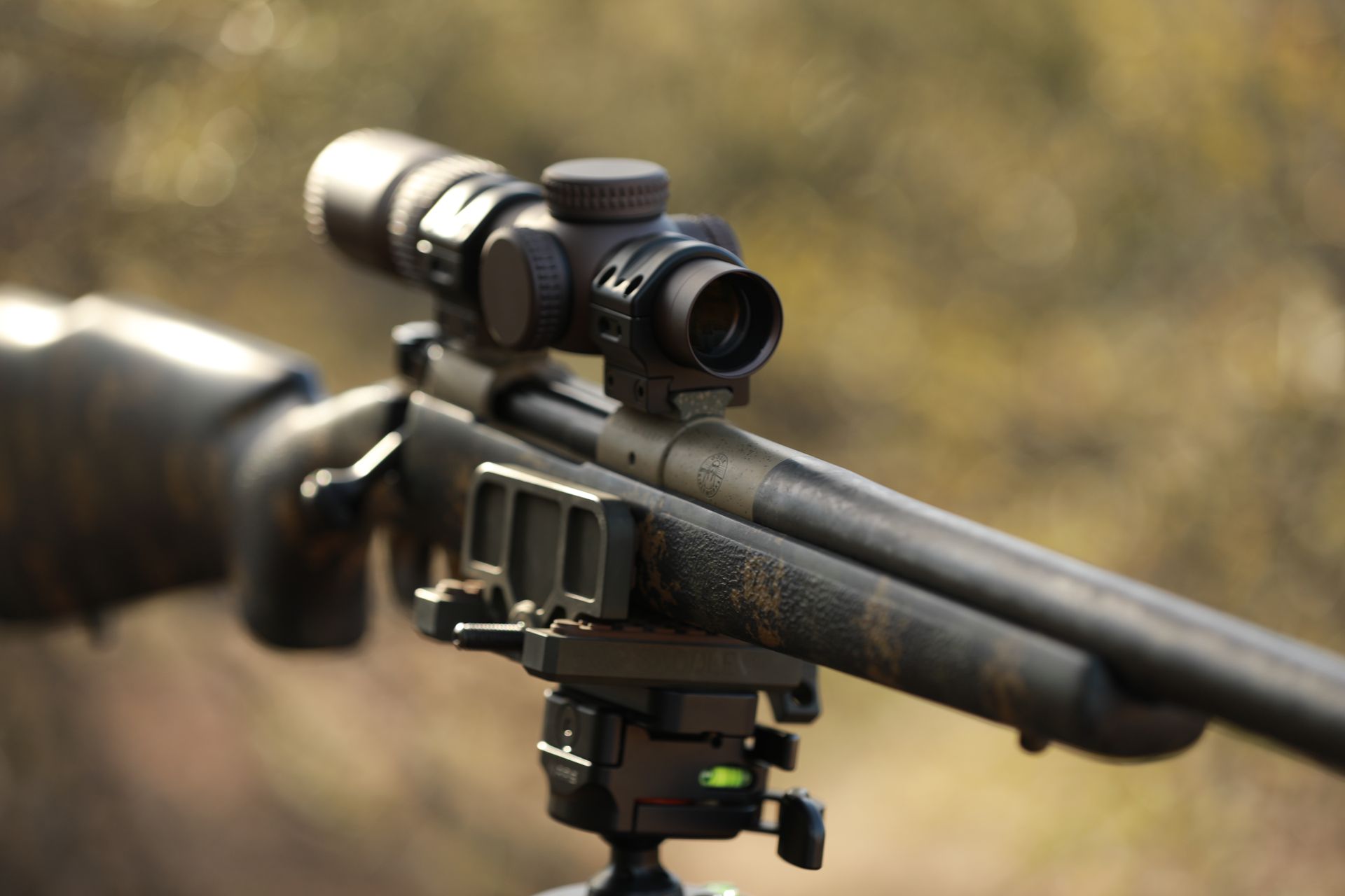 Rifle with scope mounted on a tripod, outdoors. Camouflage pattern on the stock.