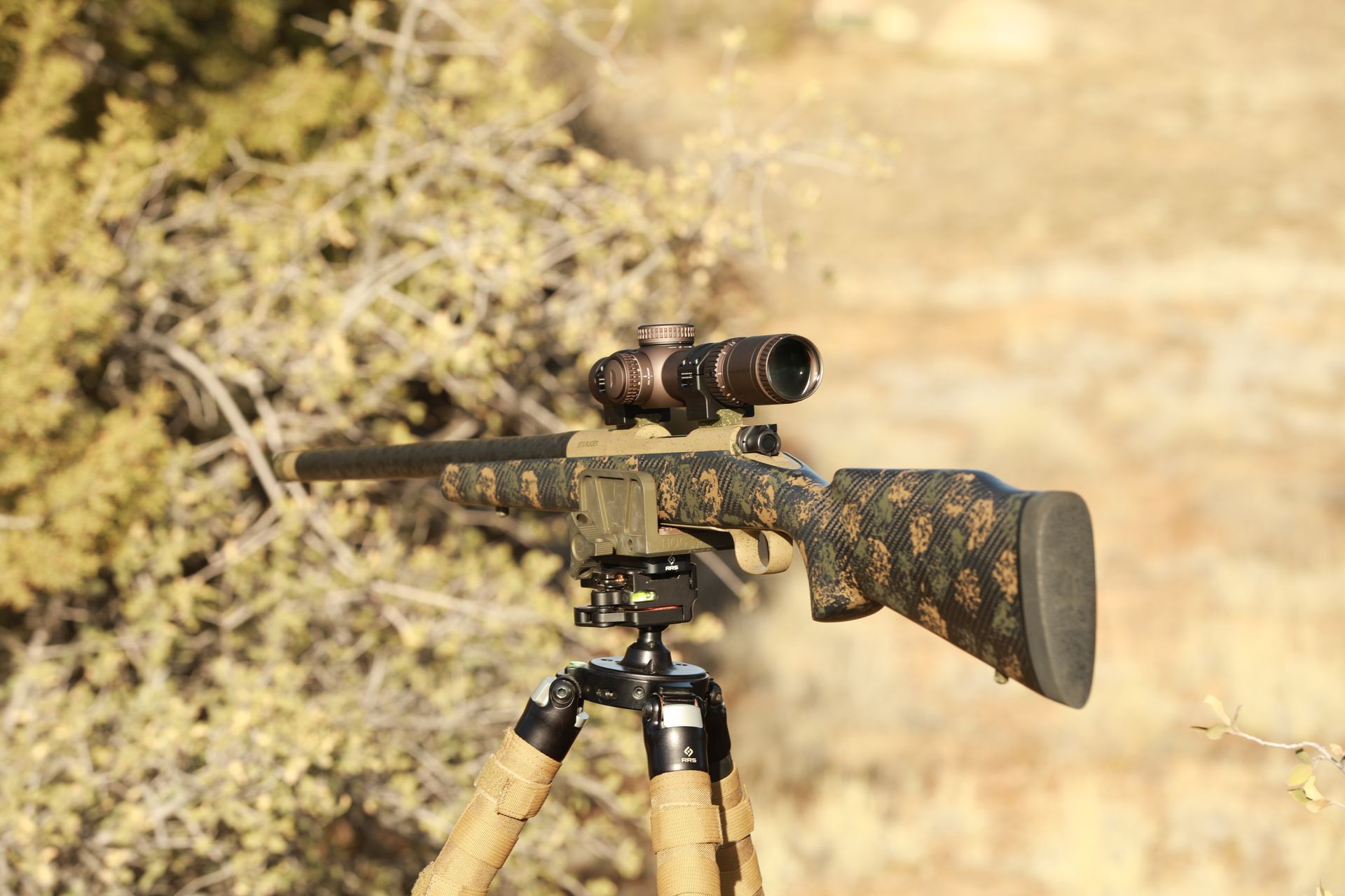 Rifle on tripod with camouflage pattern. Scope is mounted, outdoors in a tan and green environment.