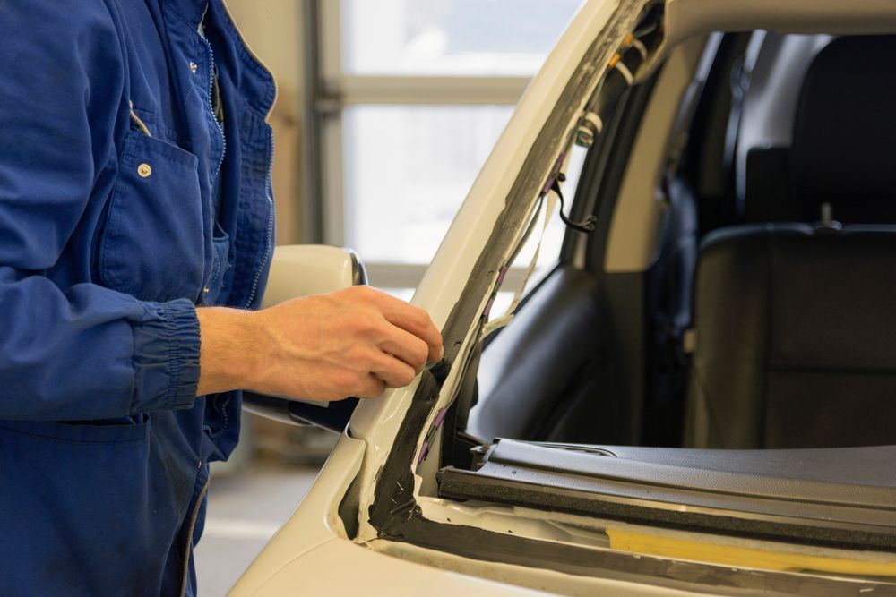 Person in a blue jacket applying sealant to a car window frame in a workshop