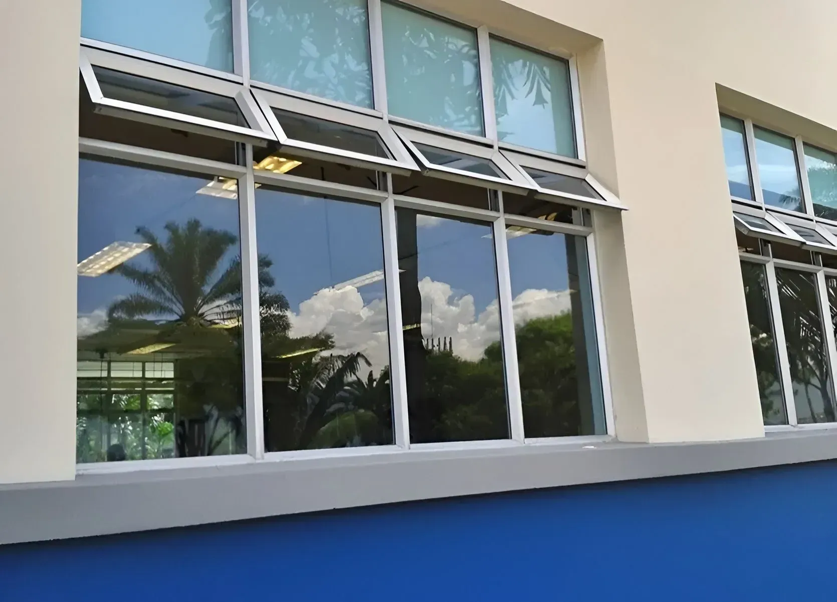 Edificio moderno con fachada de grandes ventanales y detalles en azul, que reflejan palmeras y el cielo.