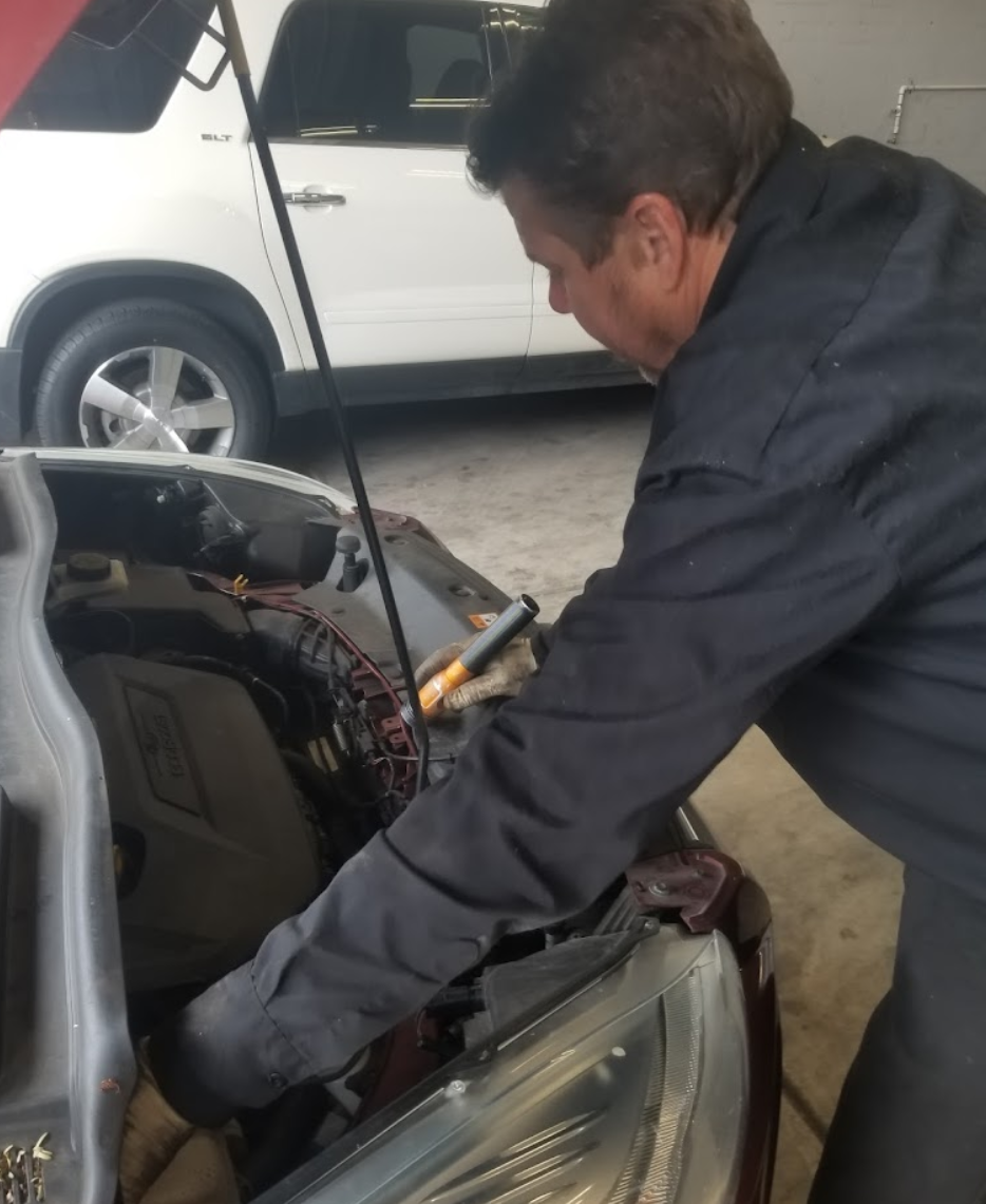 Mechanic working on a car engine in a garage, wearing a black uniform, car is maroon, white vehicle in background. | Top Notch Autocare
