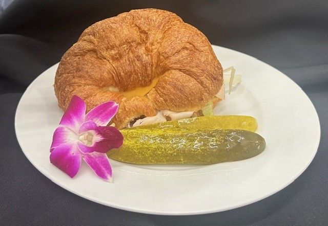 A croissant sandwich with pickles and a purple flower on a white plate.