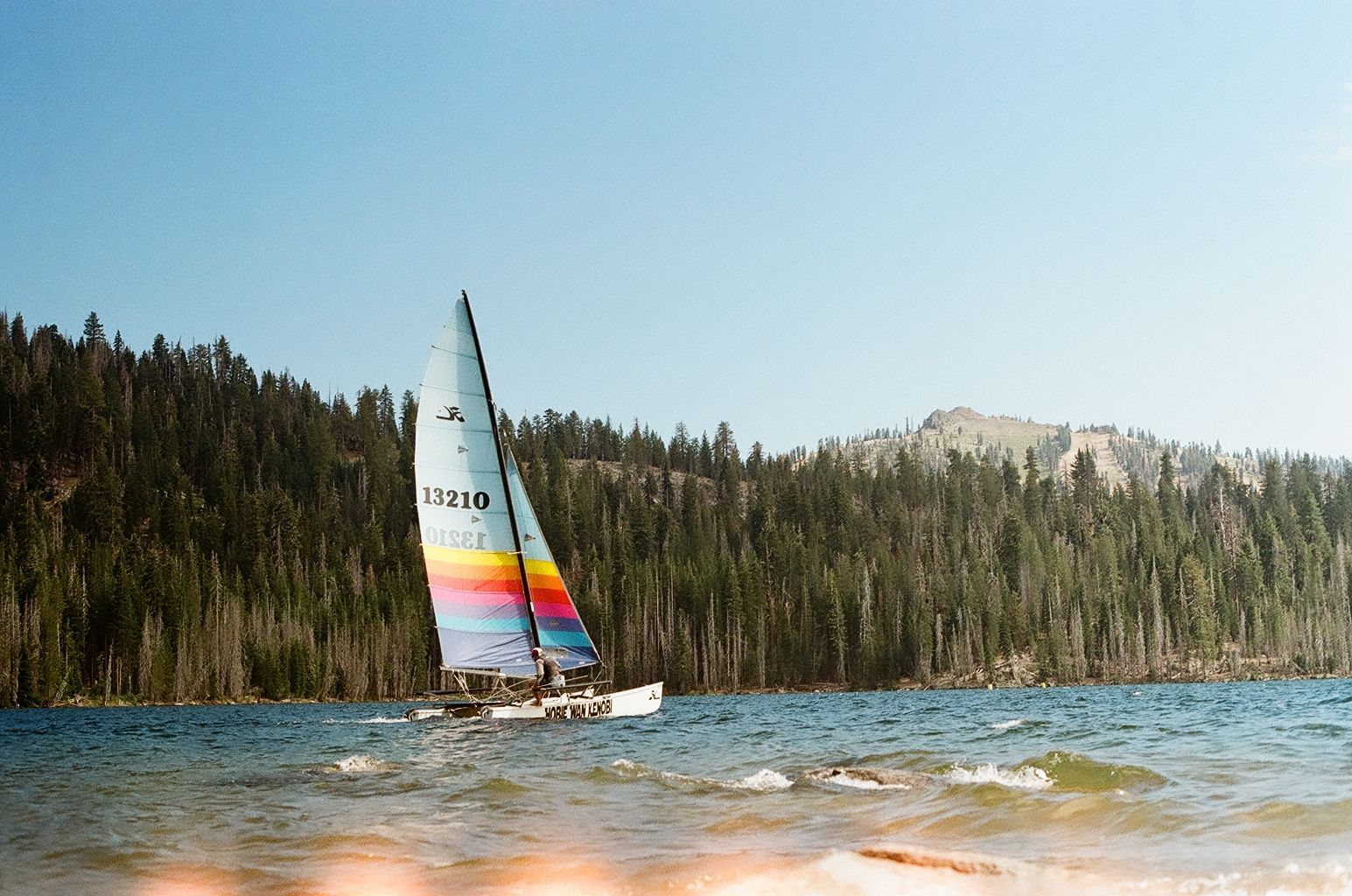 Sailboat with rainbow sail on a lake beside forested hills and snowy mountains