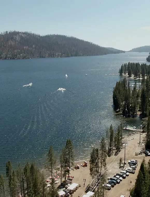 Lake with boats, beach, trees, and parked cars under a bright, sunny sky.