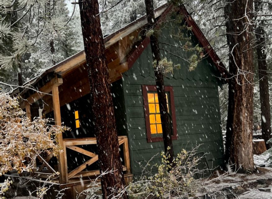 Snow-covered green cabin with lit windows in a snowy forest