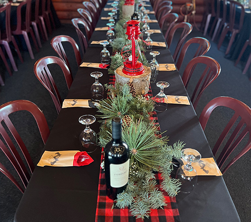 Long dining table set for an event; black tablecloth with red plaid runner, evergreen garland, wine bottle, glasses.