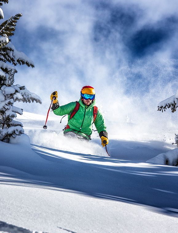 Skier carves through fresh powder, kicking up snow. Wearing green jacket, orange helmet, and blue goggles.