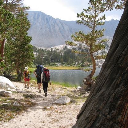 Hikers with backpacks walk a trail beside a lake, with mountains in the background.