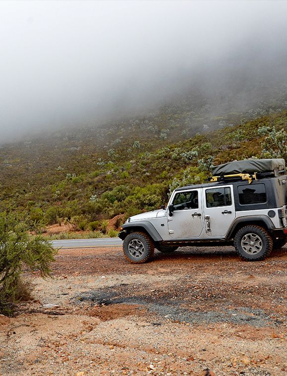 Silver Jeep on a mountain road with a rooftop tent, fog covering the hillside.