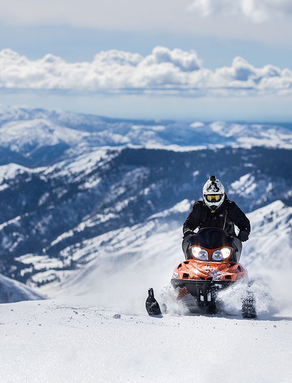 Snowmobiler rides a bright orange snowmobile on a snowy mountain, blue sky backdrop.