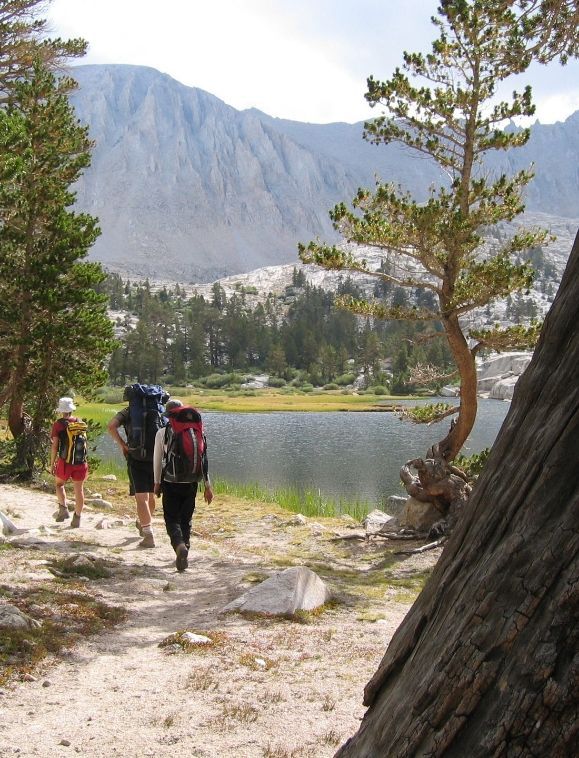 Hikers with backpacks walk along a lakeside trail, mountains in the background. Sunny day.