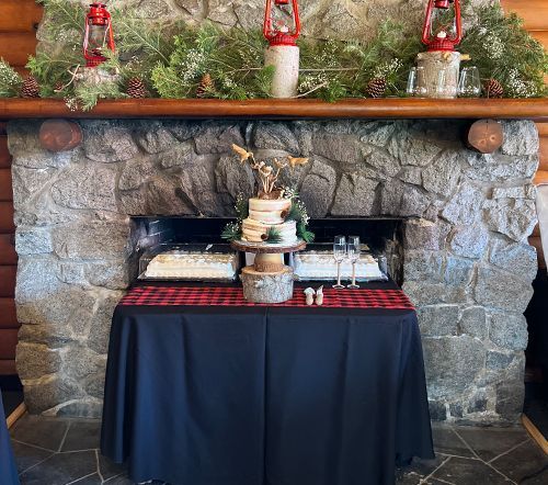 Wedding cake display in front of a stone fireplace. Cake sits on a rustic wooden stand with pine greenery and red lanterns.