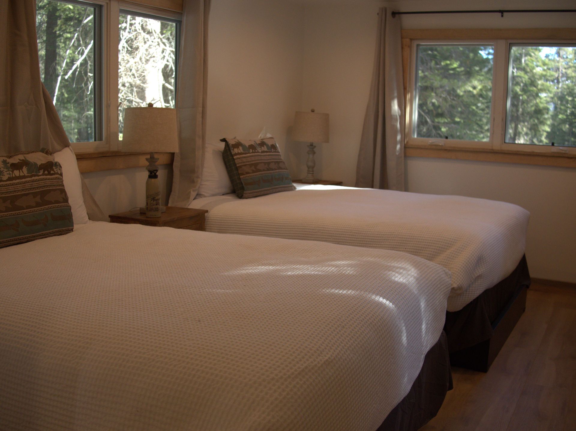 Bedroom with two white beds, beige curtains, and large windows overlooking trees.