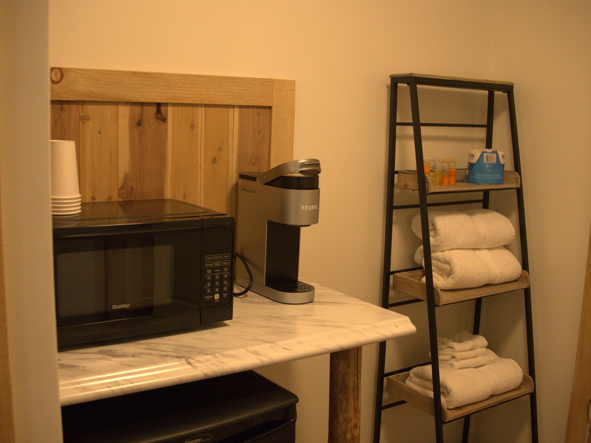 Small kitchen counter with microwave, coffee maker, and black shelf holding towels and toiletries.